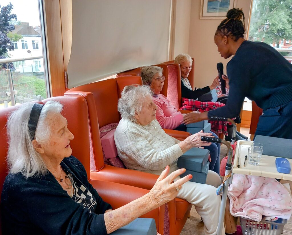 Four elderly women sit in chairs by a window, sharing good times as a carer hands a microphone to one of them. The group appears to be engaged in gentle movements and lively conversation.