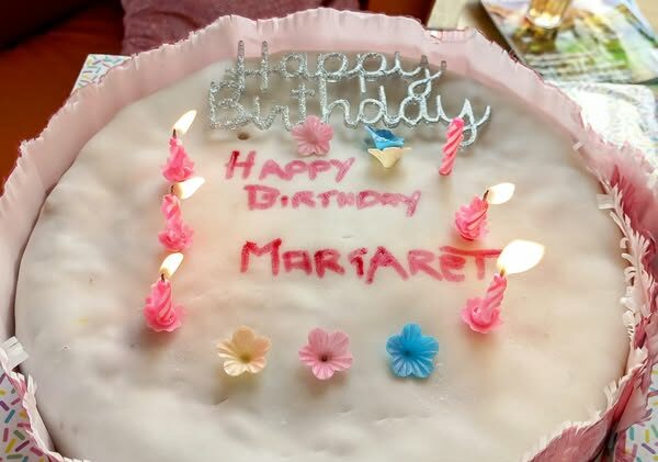 A round white birthday cake with pink icing, six lit pink candles, decorative flowers, and Happy Birthday Margaret written on top—ideal for celebrating Margaret’s birthday.