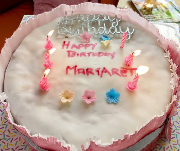 A round white birthday cake with pink icing, six lit pink candles, decorative flowers, and Happy Birthday Margaret written on top—ideal for celebrating Margaret’s birthday.