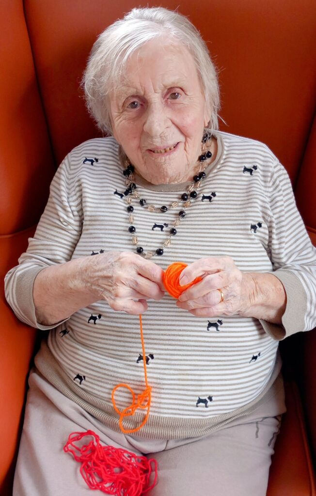 Elderly woman seated in an orange chair, holding orange yarn and smiling at the camera. She wears a striped top with small dog motifs and a beaded necklace.