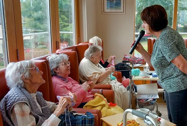 A woman holds a microphone and speaks to three elderly women seated in chairs, knitting and crocheting, in a brightly lit room with large windows.