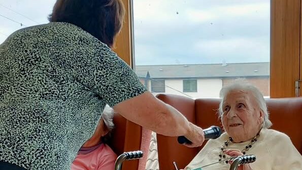 A woman holds a microphone for an elderly woman seated in a chair, who is smiling and holding a pen and paper on a tray with a glass of water and other items.
