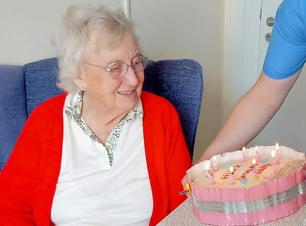 Kath, an elderly woman, sits in a chair and smiles as someone dressed in blue presents her with a frosted birthday cake topped with lit candles. Happy Birthday, Kath!.