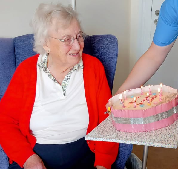 Kath, an elderly woman, sits in a chair and smiles as someone dressed in blue presents her with a frosted birthday cake topped with lit candles. Happy Birthday, Kath!.