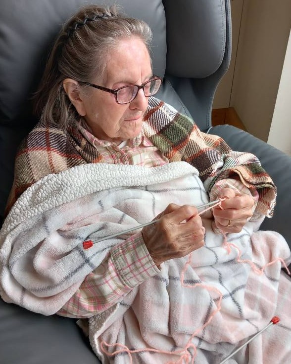 An elderly woman wearing spectacles sits in an armchair, wrapped in a checked blanket, knitting with pink wool and needles.