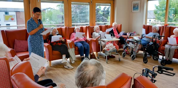 A group of older adults sit in a circle in a bright room while a woman stands reading aloud from a sheet of paper. Some participants hold papers, and mobility aids are visible.