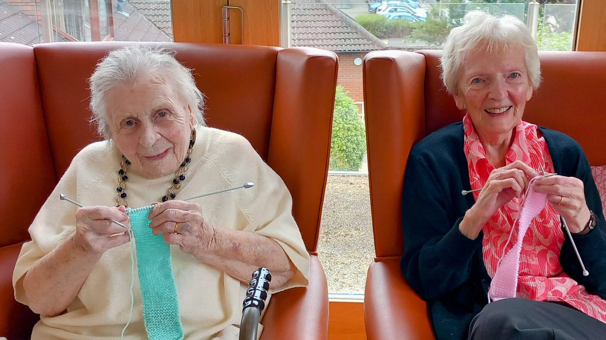 Two elderly women sit in armchairs by a window, smiling and knitting with light blue and pink wool. A walking frame is placed in front of them.