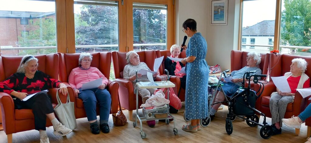 A group of elderly women sit in armchairs holding papers while a woman in a blue dress stands before them, speaking into a microphone in a brightly lit room.