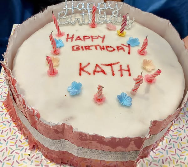 A round birthday cake with white icing, decorated with “Happy Birthday Kath” written in red, pink candles, floral decorations, and a cheerful “Happy Birthday Kath” topper.