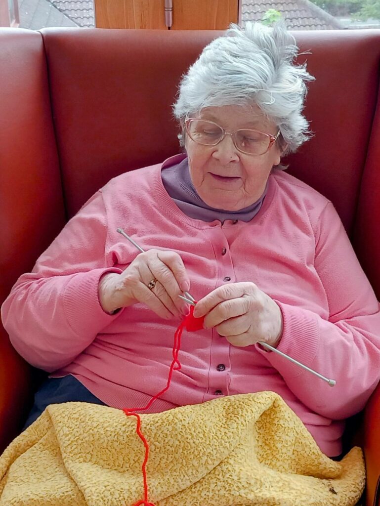 An elderly woman with grey hair and glasses sits in a red chair, knitting with red wool and metal needles, covered by a yellow blanket.