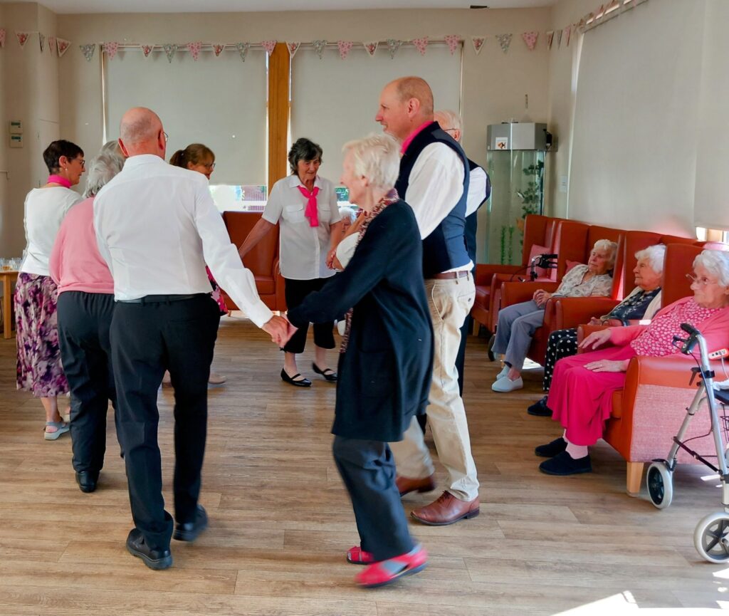 A group of elderly people enjoy a lively Dance Afternoon at Lear House, moving to the music in a brightly lit room whilst others relax on orange sofas and chairs.