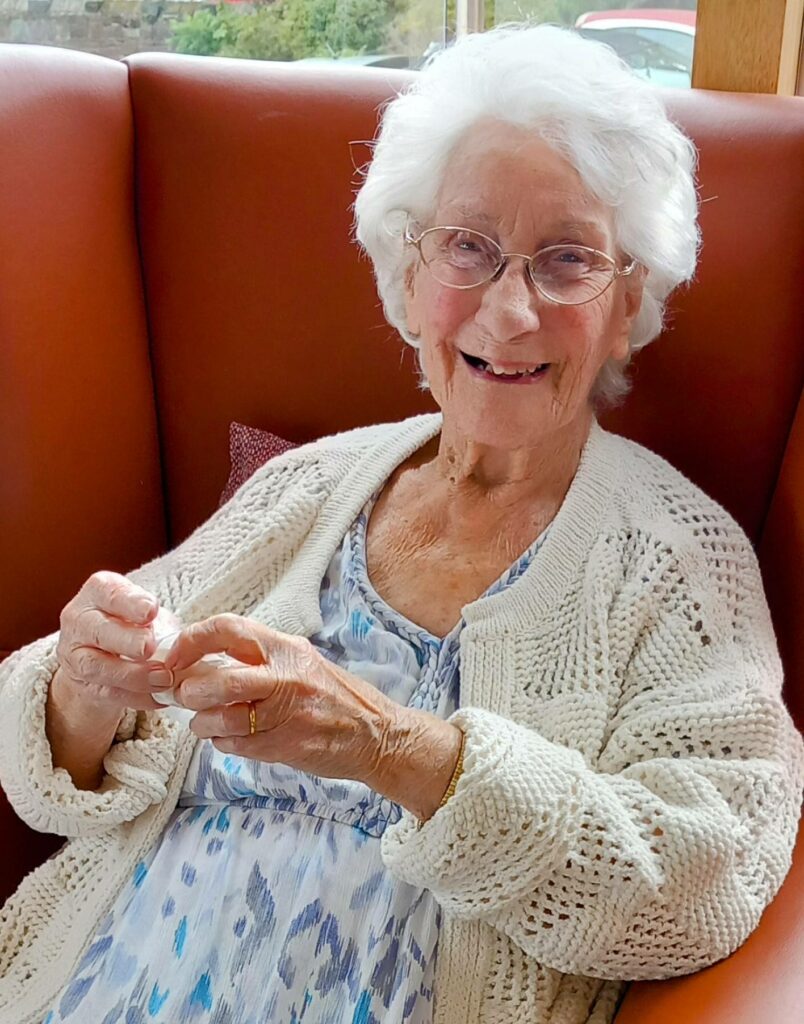 An elderly woman with white hair, spectacles, and a cream knitted cardigan smiles while seated on a brown cushioned chair.