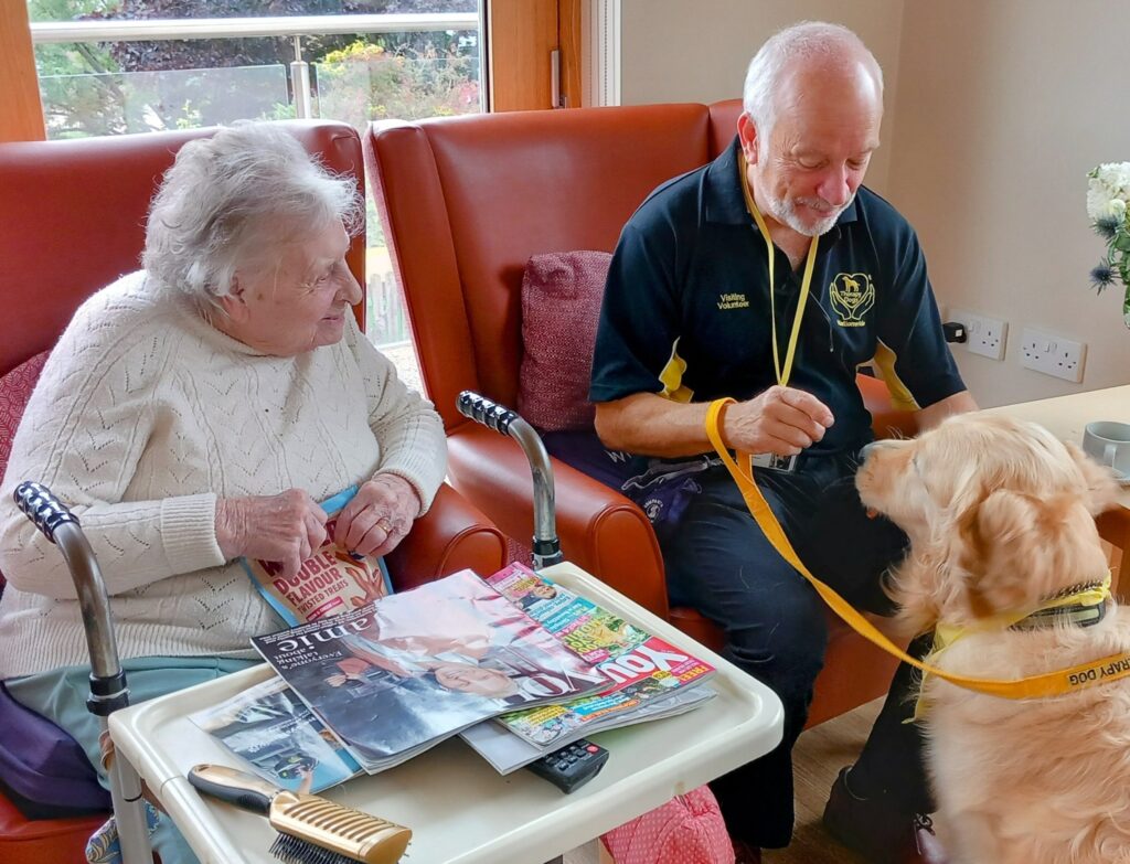 An elderly woman sits in a chair holding magazines while a man with a lanyard interacts with a therapy dog wearing a yellow vest.