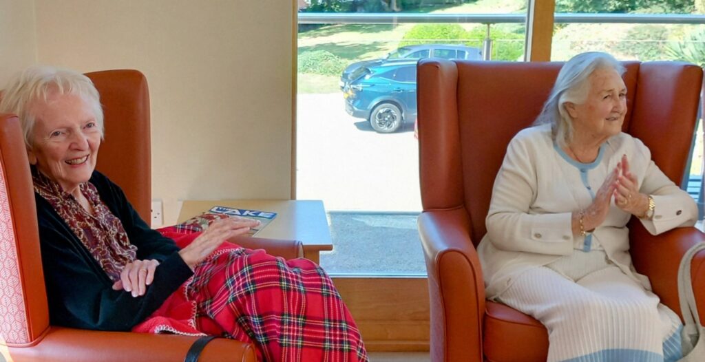Two elderly women sit in orange chairs by a window at Lear House. Sunlight pours in as one applauds, enjoying the Dance Afternoon, while the other has a red checked blanket across her lap.