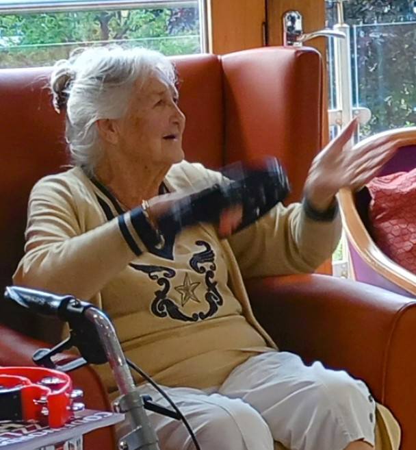 An elderly woman with grey hair sits in an armchair, gesturing with her hands, with a walking frame visible in the foreground.