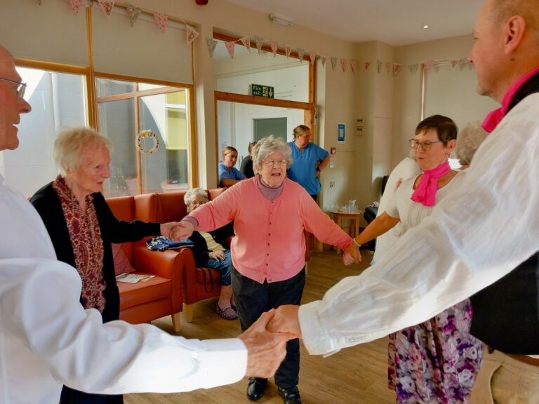 A group of older adults enjoy a Dance Afternoon at Lear House, standing in a circle holding hands and dancing in a well-lit room, while others watch from their seats in the background.