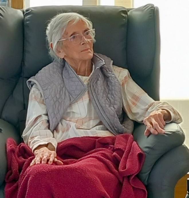 Elderly woman with short white hair and spectacles sits in a reclining chair at Lear House, wearing a checked shirt, quilted gilet, and covered with a red blanket during the Dance Afternoon event.