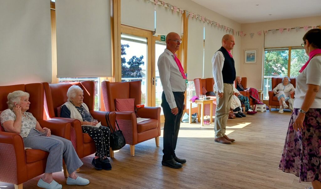 At Lear House, a group of elderly people sit in armchairs while three adults wearing pink scarves host a lively afternoon of dancing. Sunlight pours through large windows into the bright, cheerful room.