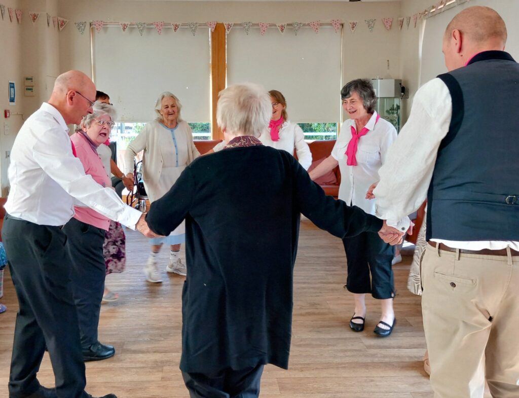 Older adults stand in a circle holding hands at the Lear House, enjoying a lively Dance Afternoon together indoors.