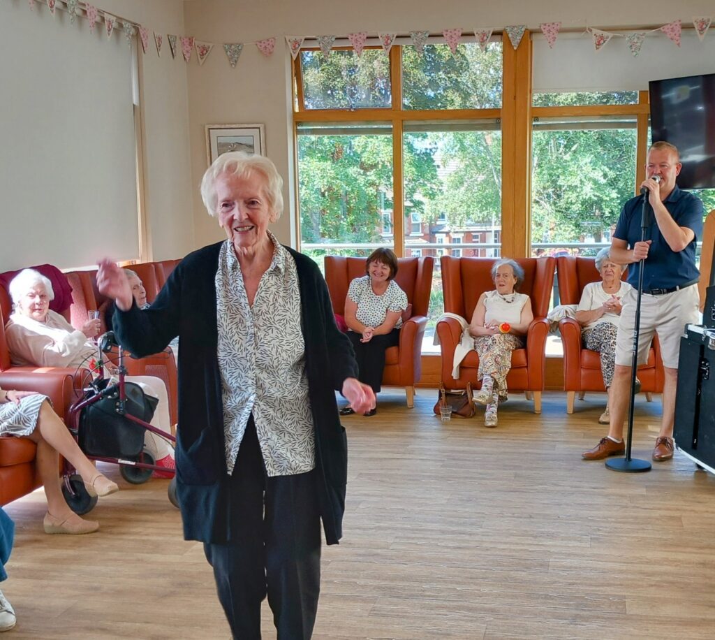 An elderly woman smiles and dances in a care home lounge, sharing moments of companionship as other residents watch and a man sings brilliant music into a microphone.