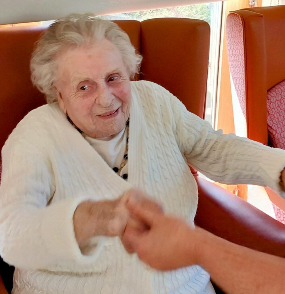 Elderly woman in a white jumper sits in an orange chair at Lear House, smiling and holding hands with another person during Dance Afternoon.