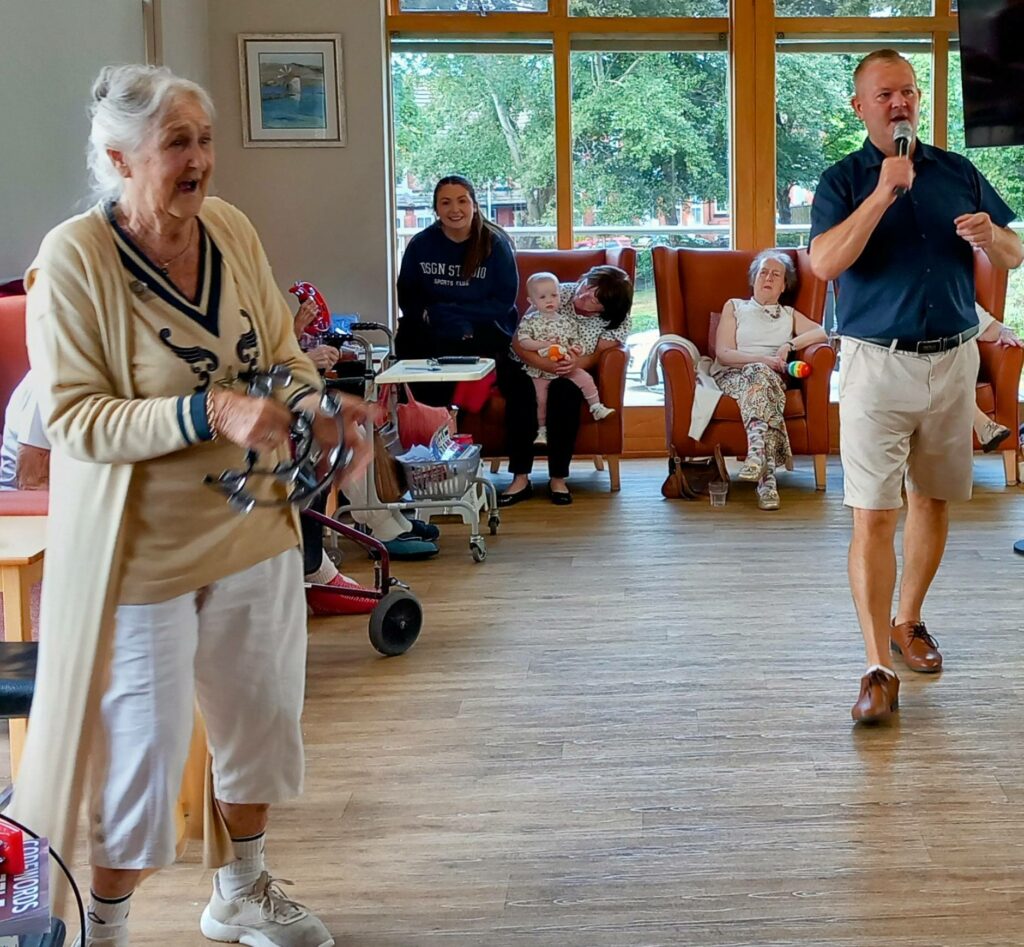 An elderly woman plays a tambourine while a man sings into a microphone; several people sit and watch, enjoying wonderful music and camaraderie in a bright, communal room.