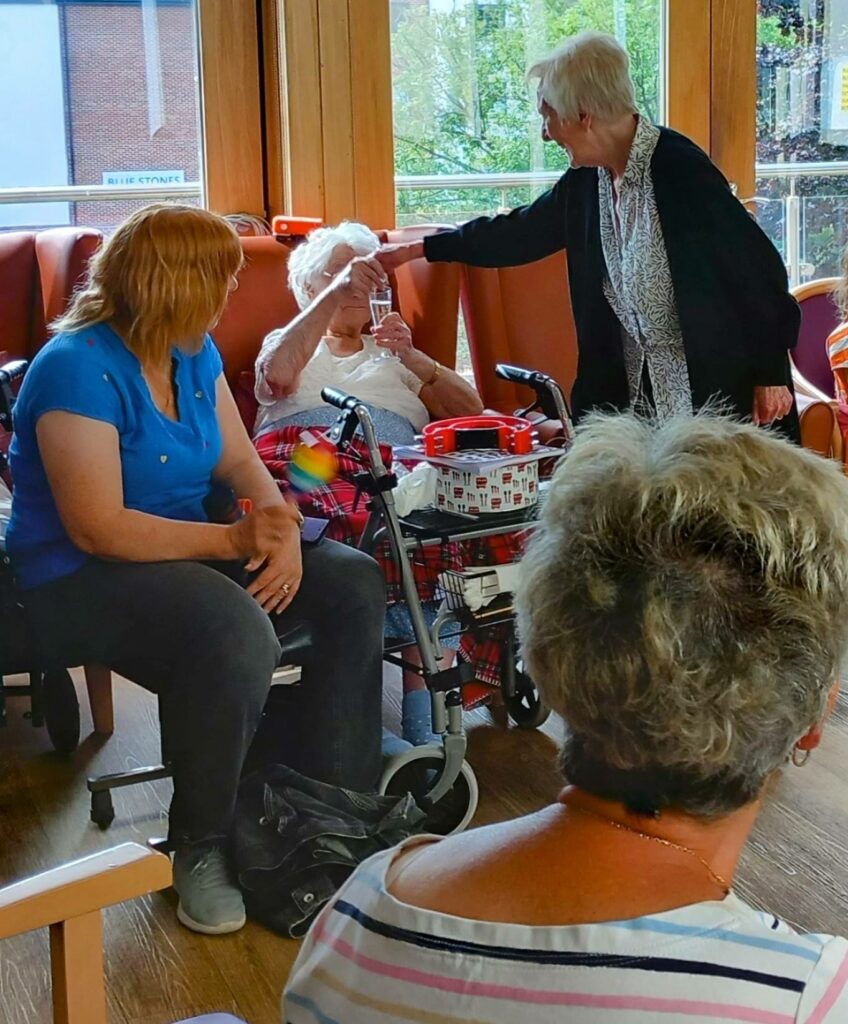 Four women celebrate friendship in a sunlit room—one in a wheelchair holding a glass of fizz, another reaching out warmly. Presents and decorations contribute to the fabulous atmosphere.