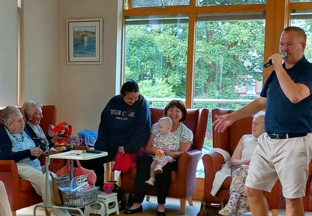A group of older adults and a baby sit together indoors, enjoying marvellous music as a man stands before them singing into a microphone, celebrating friendship across generations.