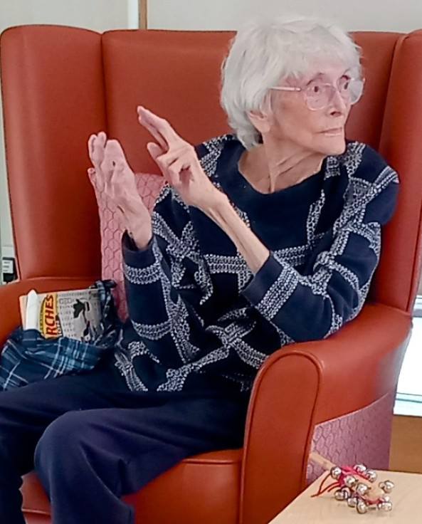 An elderly woman with white hair and spectacles sits in an armchair, dressed in a navy patterned jumper. A magazine and tartan bag are by her side as she gestures animatedly, reminiscing about a joyful afternoon with The 64 N’Ukes ukulele band.