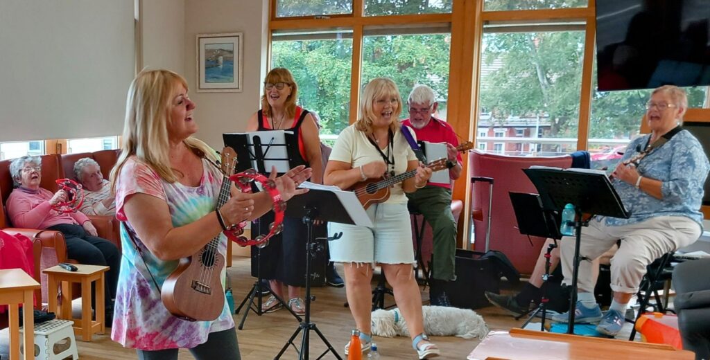 The 64 N’Ukes play musical instruments and sing together in a brightly lit room with large windows, creating a joyful afternoon as some seated people watch and a dog lies on the floor.