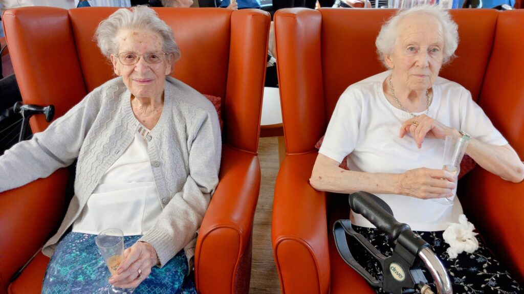 Two elderly women sit beside each other in orange armchairs, each holding a drink. One wears a grey cardigan, the other a white top—a touching farewell gathering for Sarah. A mobility aid is visible in the foreground.