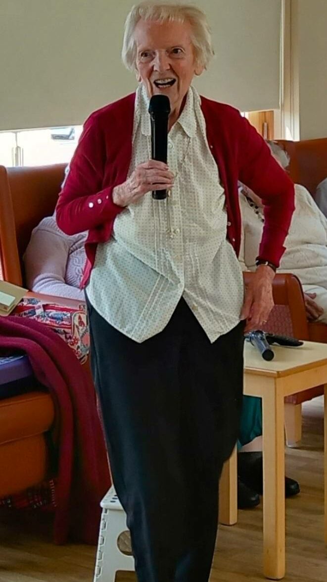 An elderly woman stands indoors, holding a microphone and smiling at Sarah’s farewell after 28 years’ dedication. She wears a red cardigan, light patterned blouse and dark trousers; others are seated in the background.