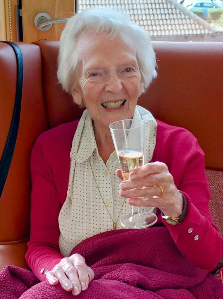 Elderly woman with white hair smiles while holding a glass of champagne, seated on a brown chair with a maroon blanket over her lap, celebrating Sarah’s leaving do and 28 years’ service before retirement.