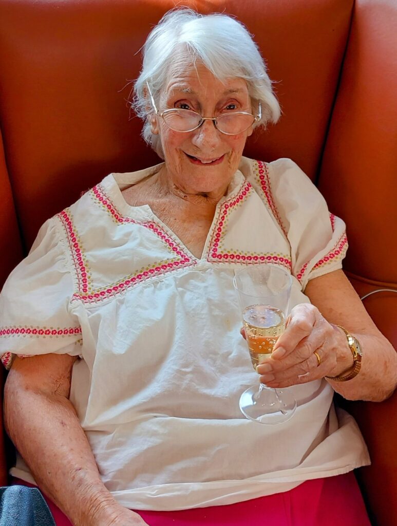 Elderly woman with white hair and spectacles sits in an armchair, smiling and holding a glass of fizzy drink, celebrating Sarah’s farewell with warmth and dedication.