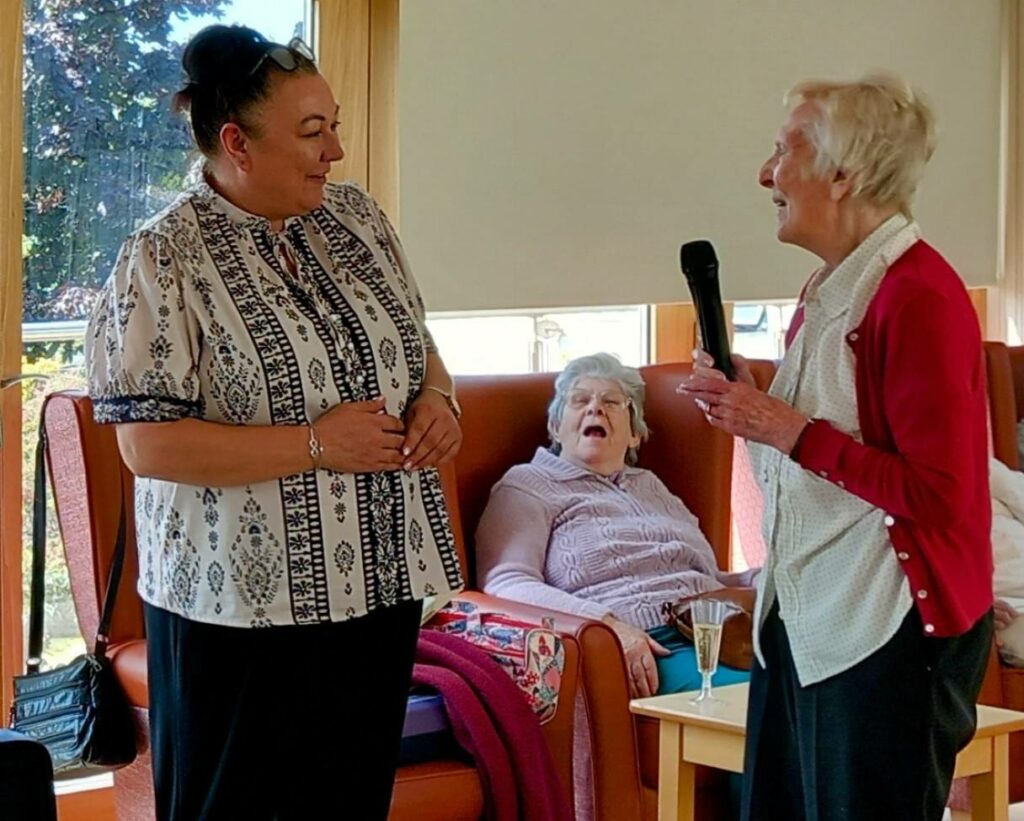 An older woman demonstrates her dedication as she holds a microphone and speaks to another woman named Sarah, while a third woman sits on a chair in the background with her mouth open.