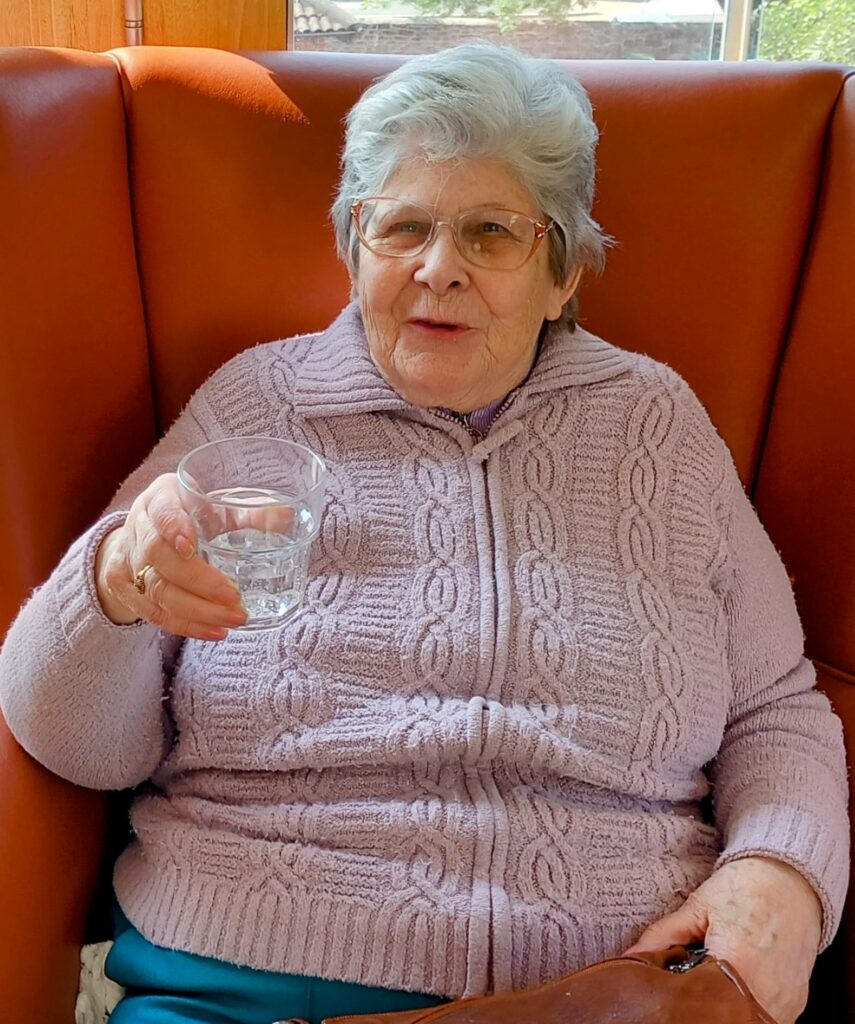 Sarah, an elderly woman with grey hair and glasses, sits on an orange chair in a light purple jumper, holding a glass of water and reflecting her dedication over 28 years as she looks at the camera.