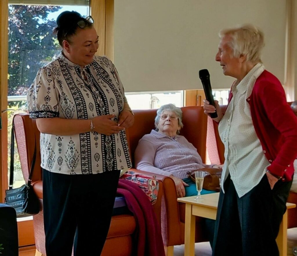 Two women stand and chat, one holding a microphone, while an elderly woman—recognised for her 28 years of dedication—sits in an armchair behind them in a brightly lit room.