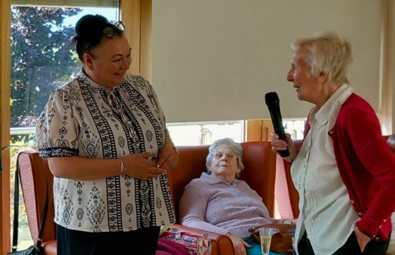 Two women stand and chat, one holding a microphone, while an elderly woman—recognised for her 28 years of dedication—sits in an armchair behind them in a brightly lit room.