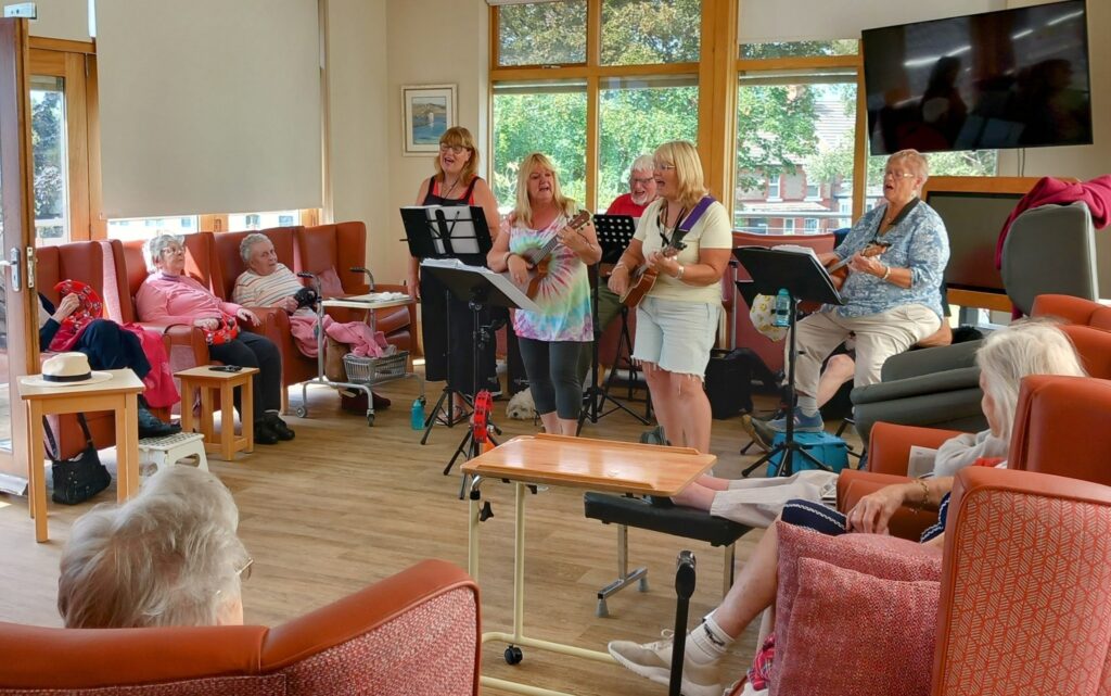 A small group of women from The 64 N’Ukes play musical instruments and sing in a lounge, creating a joyful afternoon as elderly people sit and watch from armchairs.