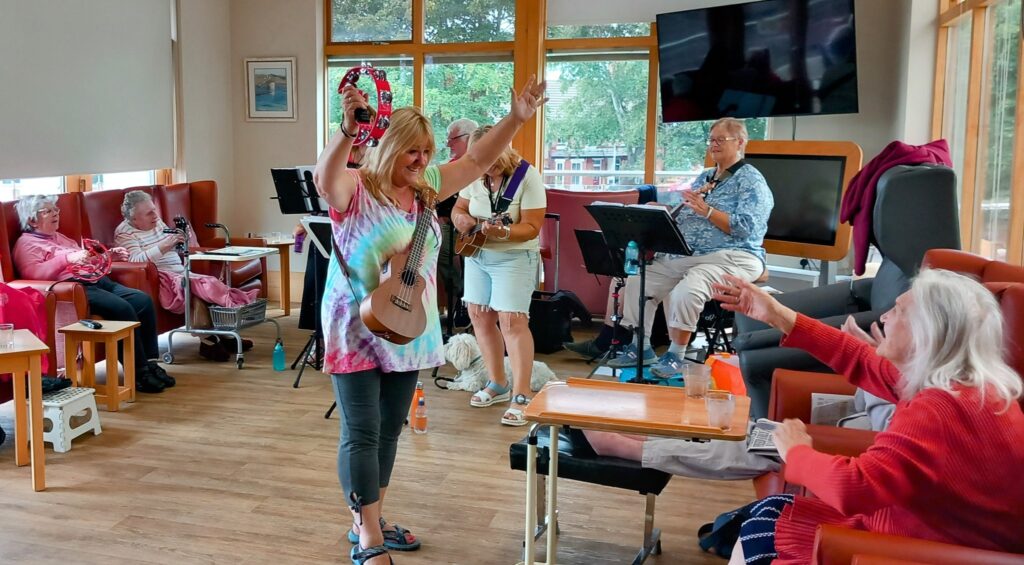 A woman holding a ukulele and tambourine engages with seated pensioners in a lounge as The 64 N’Ukes Ukulele Band performs music in the background, creating a cheerful afternoon for all.