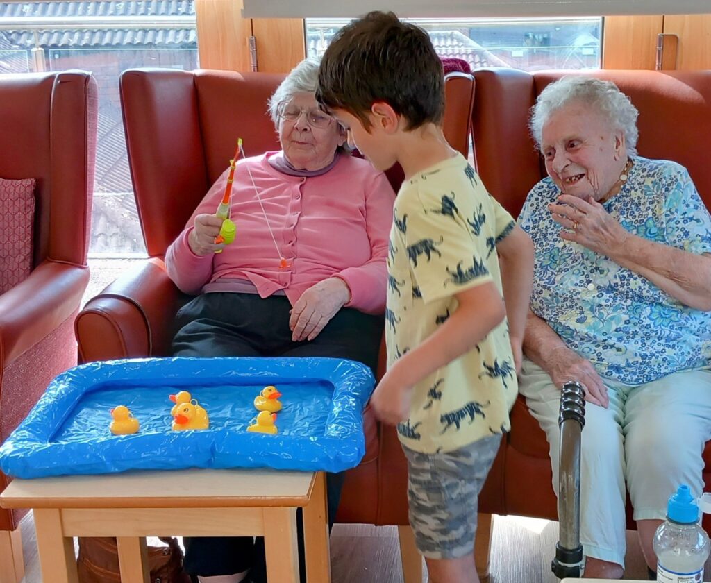 A young boy plays a fishing game with toy ducks in a small paddling pool as two elderly women watch from their armchairs and smile.