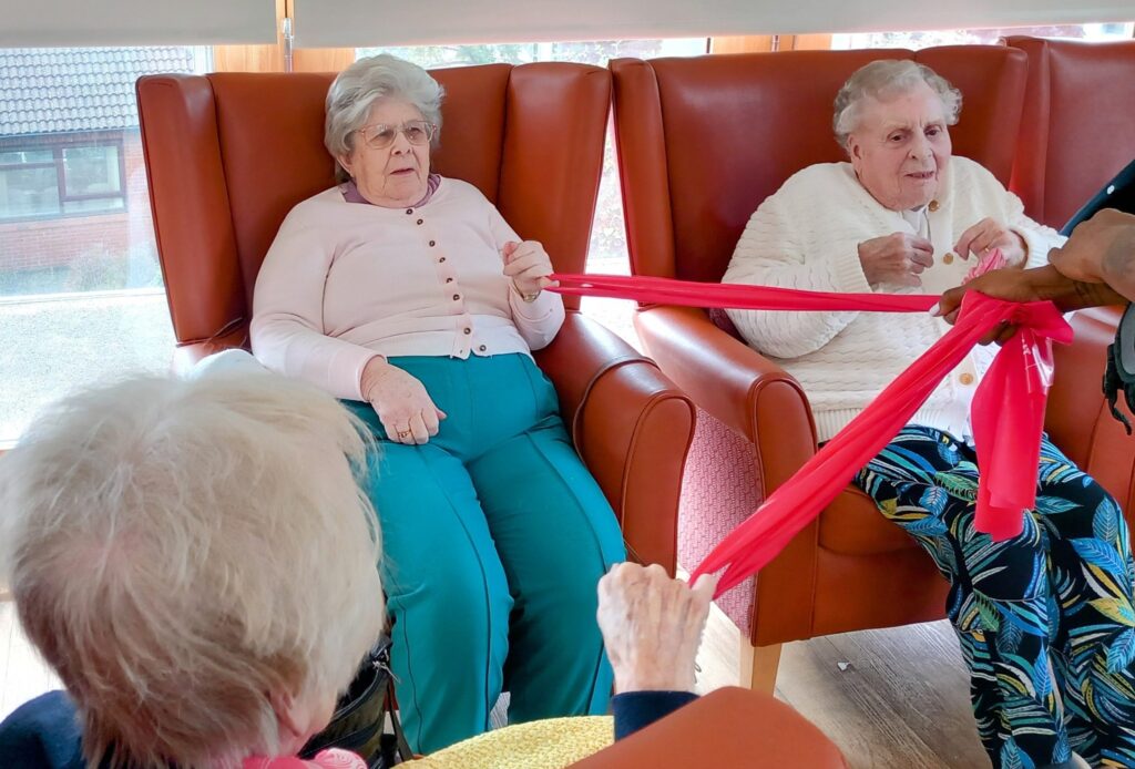 Three elderly women seated in armchairs take part in a group exercise, stretching a long red resistance band held by someone standing close by.