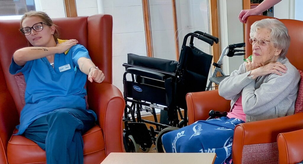 A healthcare worker and an elderly woman sit in armchairs, each touching their own shoulder, with a wheelchair visible in the background.