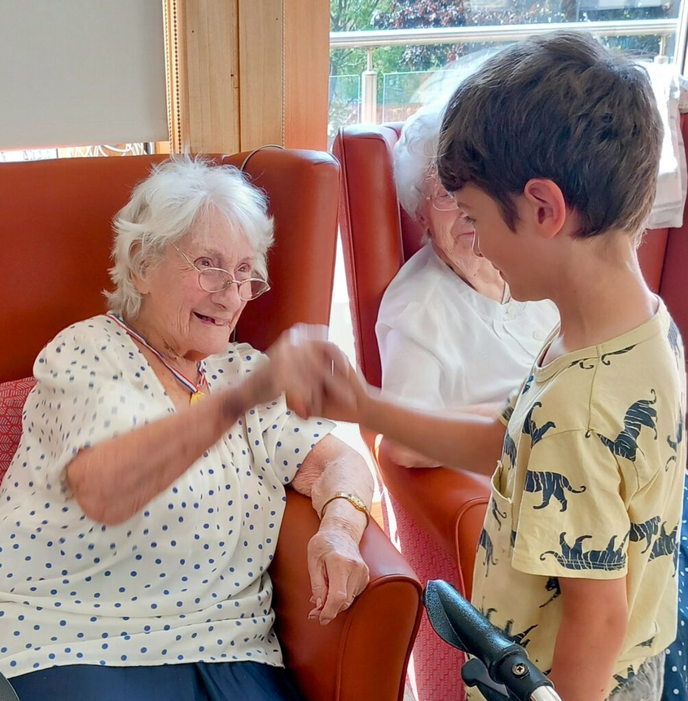 An elderly woman sitting in a chair smiles and gives a fist bump to a young boy standing before her, with another elderly woman seated nearby.