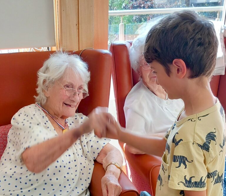 An elderly woman sitting in a chair smiles and gives a fist bump to a young boy standing before her, with another elderly woman seated nearby.