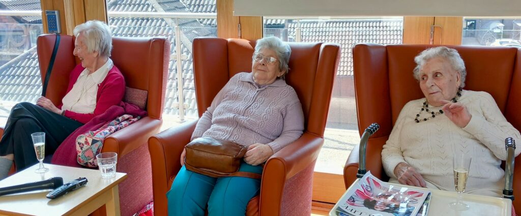 Three elderly women sit in armchairs by a window, sharing drinks and magazines as they celebrate Sarah’s farewell and her 28 years of dedication to retirement living.