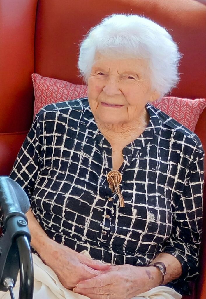 Elderly woman with white hair, known as Sarah, sits in a red armchair wearing a black and white patterned top, smiling softly with her hands in her lap—a testament to 28 years of dedication.