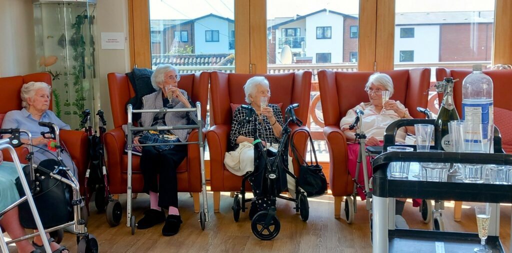 Four elderly women, including Sarah, sit in armchairs with walking frames before them, enjoying drinks in a bright room with large windows—a testament to 28 years of devotion and friendship. A drinks trolley is visible at the front.