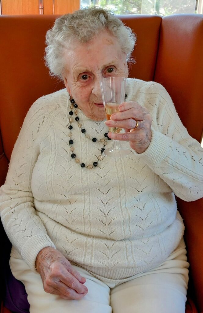 Elderly woman in a white jumper seated on an orange chair, raising a glass with a drink and wearing a beaded necklace, celebrating Sarah’s 28 years of dedication.