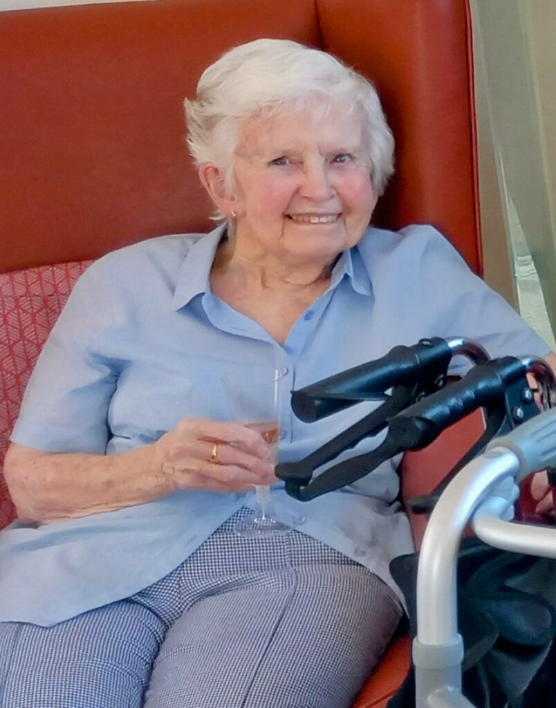 Elderly woman with short white hair sits on a red chair, smiling and holding a drink, her walking frame in front of her—celebrating Sarahs farewell after 28 years dedication before retiring.
